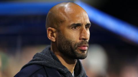 Lee Grant looks into the distance with a focused look on his face. he has black stubble and wear a dark grey knitted hooded cardigan with a navy jacket