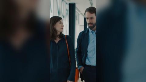 Jodie Whittaker and Joseph Fiennes walk down a corridor wearing navy tops.
