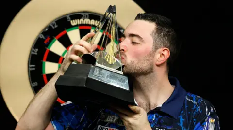 Luke Humphries celebrates with the Premier League Darts trophy after winning the final match against Luke Littler