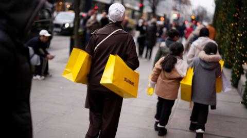 Shoppers on Oxford Street walk along the pavement holding bright yellow Selfridges bags with Christmas trees on the right