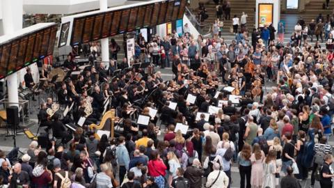 The view down towards the departure boards at a railway station. Members of an orchestra play in front of the signs, with a large number of people surrounding them.