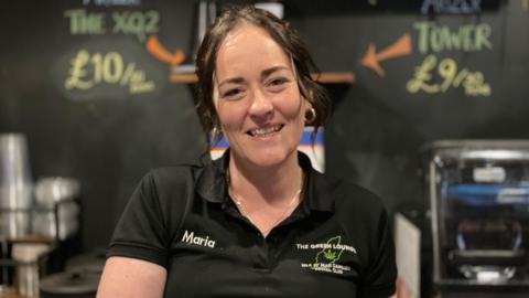 Maria Leonard is wearing a black polo shirt with the Green Lounge written as a logo on the right with a green outline of the Isle of Man. She has dark hair tied up with wavy tendrils 