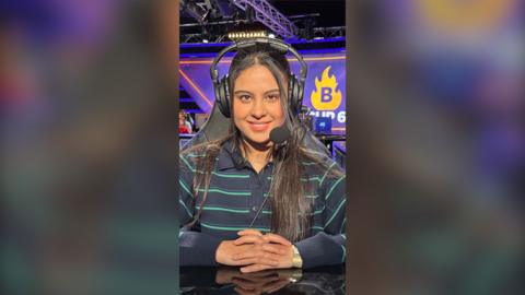 A young woman is sitting in a computer chair, in an e-sport venue. She is looking directly into the camera, smiling. She is wearing a striped shirt and a headset.