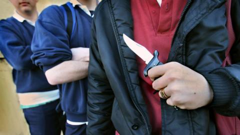 Three young men in the picture. Two of them have their arms crossed and the other is holding a small knife in his hand. 