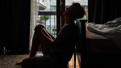 a young man sits holding his knees with his head leaning back in a dark bedroom with light from glass doors leading on to a small balcony