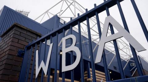 Shot of the outside of The Hawthorns stadium looking through a side gate with the club's acronym WBA on the front
