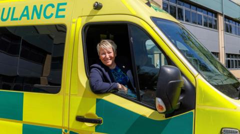MP Rachel Taylor is sitting at the wheel of an ambulance which is liveried in yellow and green. It is parked outside an office building.