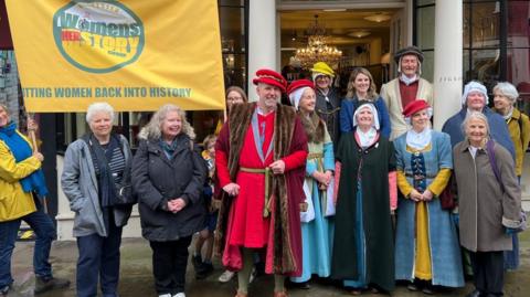A group of people, some in period clothing, stand beneath a yellow banner outside a shopfront with decorative pillars.