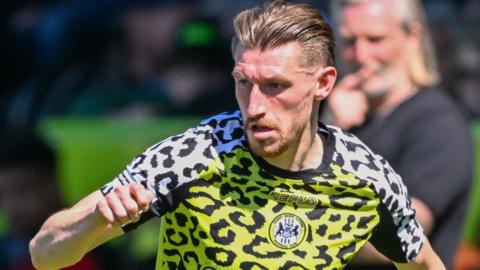 Tom Knowles is wearing a Forest Green home kit which has a yellow leopard print body with white leopard print arms while manager Robbie Savage looks on in the background.