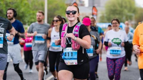 A runner wearing sunglasses, black t-shirt, black shorts and a pink hydration vest smiles into the camera as she takes part in the Grimsby 10K. A number of other runners are visible in the background.