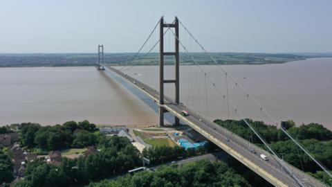 Aerial view of the Humber Bridge showing the bridge spanning the muddy water with vehicles crossing.
