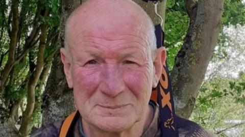 Stephen Woodward is looking at the camera in a close-up shot of his face. He is balding and is standing outside with trees behind him.