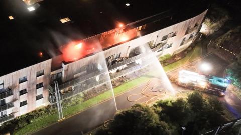 An aerial shot of water being sprayed towards the top floor of a flat with a fire engine at the base of the building