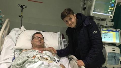 Jack Rondel pictured with his father Richard in hospital. Richard is lying in bed, while Jack is stood bedside in a black coat. They are both smiling, Richard is wearing black glasses. hospital equipment is visible in the background.