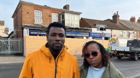 Simba Chitsatso, left, and his wife Milly. Simba is wearing a bright yellow rain jacket and Milly is wearing an olive green jacket and sunglasses. Both are looking at the camera and the burnt remains of the shop and their flat are in the background. A plywood wall has been erected around the shop frontage and parts of the signage are completely melted.