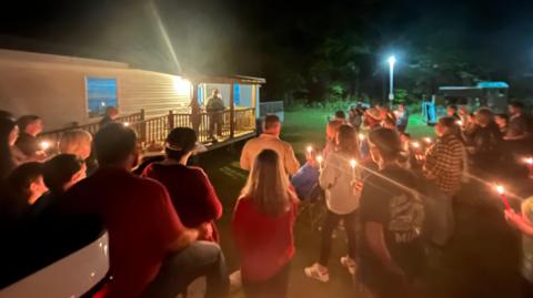 A crowd holds candles and listens to a speaker outside a small building