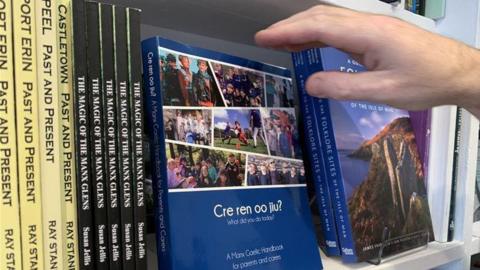 The Cre Ren Oo Jiu? book sits on a bookshelf. It is blue and has pictures on children doing activities, such sa playing sport. There are other books on folklore and glens that sit next to it.