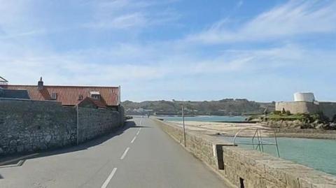 Google Street View image of Route de Rocquaine in Guernsey with the sea and a fort on the right-hand side and a large stone wall and a residential property on the left.