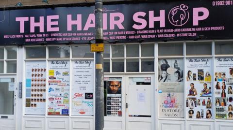 The front of a large shop with panelled windows. Numerous posters of hair and hair products can be seen advertised on the front.