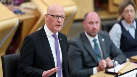 a bald man with glasses holds out his right hand as he is answering a question from across the chamber. He is wearing a dark jackeet, white shirt and purple tie. There is one male and one female colleague seated next to him