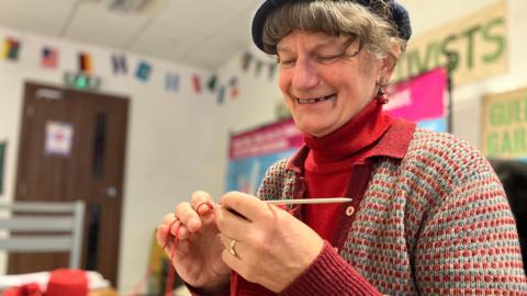 A woman is knitting with red wool, and smiling. She sits inside a room with banners and flags on the wall.