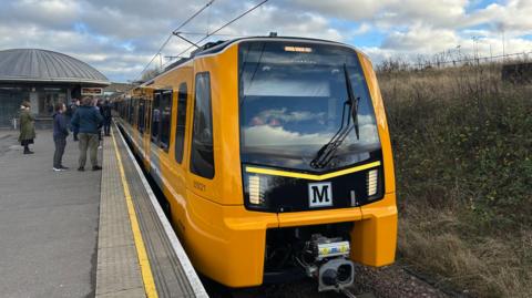 A new model of metro train at Pelaw station. The carriage is yellow with the 'M' metro logo on the front. People are waiting to get on the train.
