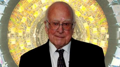 Prof Higgs is looking at the camera. He is bald apart from some white tufts of hair at the sides of his head. He is wearing glasses and a dark suit and tie.There is a bright yellow and green circular background