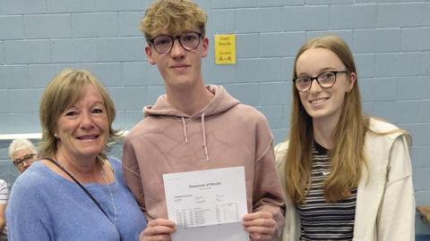 Beckett stands between his mum and sister holding his results, in a school hall.