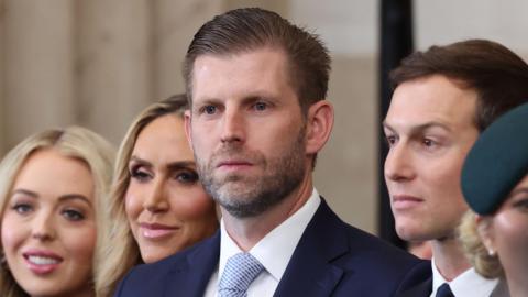 Eric Trump, visible from the shoulders up. He is wearing a blue blazer, a white shirt and a light blue tie and is surrounded by family.