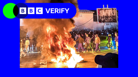Huge fire and plume of smoke in the centre of the image with crowds of masked people surrounding it during Tehran protest 