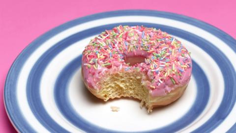 Frosted pink doughnut on a plate which has a blue and white circular pattern. A bite has been taken out of the doughnut.
