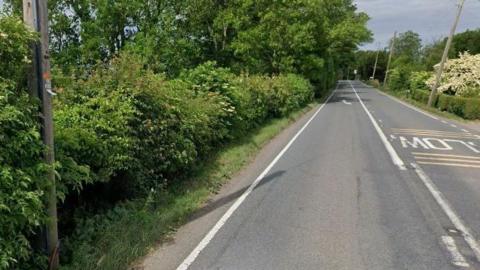 A stretch of an A-road with a hedge on one side and trees in the distance. 
