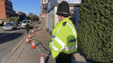 A police officer wearing a high viz yellow coat and tall black police helmet stands beside a row of small orange cones with police tape marking out the crime scene. 