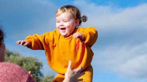 A little girl in an orange jumper and trousers is playing with her mum outside on a sunny day. The mum has her back to the camera and is opening her arms to catch the girl. 