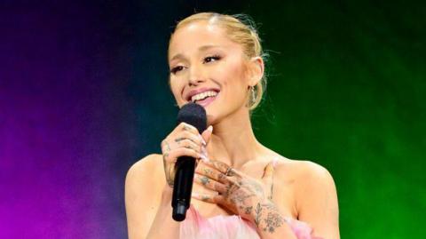 Ariana Grande holding a black microphone smiling and singing
