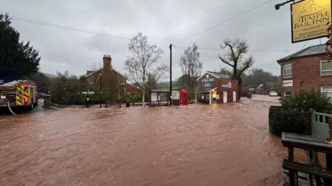 Dark orange floodwater running through a village. The water has covered the road and is sweeping through the street. There is a pub on the right and a bus stop, phone box and fish and chip shop that are all flooded. A fire engine is on the left trying to pump out the water.