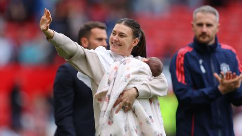 Hannah Blundell smiles and waves while holding her baby on a football pitch