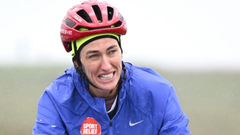 Jill Scott on the road during day four of the Coming Home Challenge. She is grimacing while wearing a purple raincoat and a red cycling helmet which are wet from the rain.