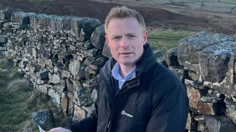 Robbie Moore MP standing in front of a stone wall