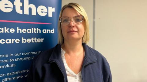 A woman standing in front of a blue promotional board with blonde hair and is wearing glasses with a white shirt and blue fleece zip-up top.