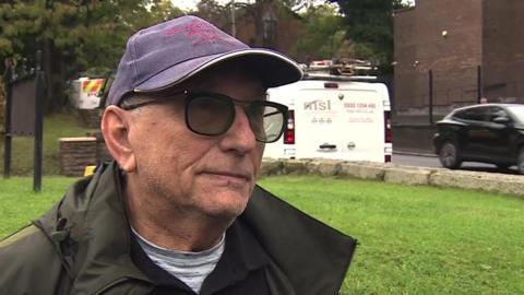 An older man stands looking at someone off-camera. He is standing outside and wearing a cap, tinted glasses and a dark green jacket. 