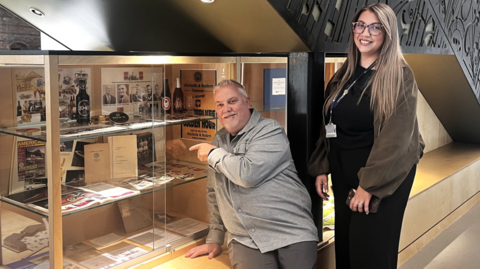 Stuart points to brewery artifacts on display in a glass cabinet in the university building with his daughter stood beside him. Stuart is wearing grey trousers and a grey top. Leigh is dressed in black with a brown top and has long hair and glasses on.