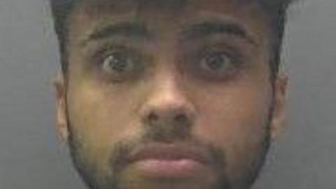 Mugshot of Isaiah Khan. He is brown and has dark hair [ short back and sides and long on top]. He has a dark beard and moustache, which is groomed close to his skin. 