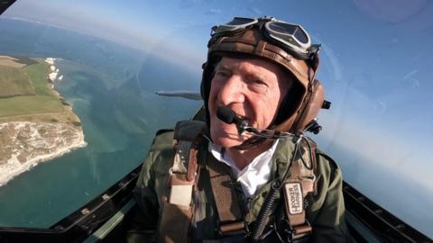 Brian Fox in the spitfire in the sky. He is wearing a helmet with a coms system, and a green jumpsuit. They are flying above the sea, with cliffs on the left hand side.