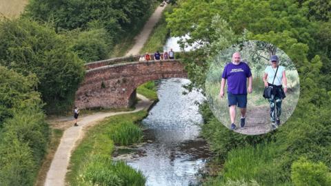 A group of people standing on a bridge over a river, with one person standing on a path beside the river