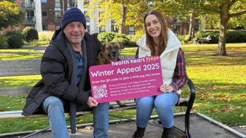 A man wearing a dark coat, blue jeans and a wooly hat sits on a park bench next to a young woman with long dark hair, a white coat and blue jeans. They are holding a board with the name of a charity on it, while a Staffordshire terrier sits between them peering over the top of the board.