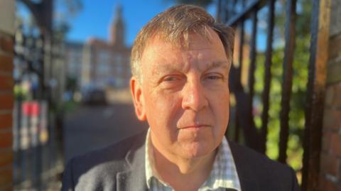 Sir John Whittingdale, standing outside the metal gates of Maldon hospital. He is wearing a green checked shirt and a dark coloured suit.