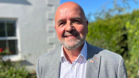 A bald man smiling at the camera wearing a grey suit jacket on top of a white shirt, with a Guernsey flag pin on his lapel. The background of a white building to the left and green bushes to right are blurred. Clear blue skies.