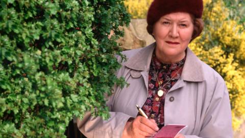 A woman standing just behind a bush taking notes on a notepad.
