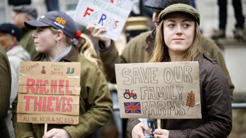 Two women at the protest holding signs. One says "save our family farms" and the other "Rachel Theives All the Keir and No Idea" 